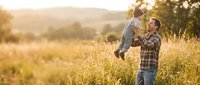 Happy father lifting baby son in golden sunset field, family bonding moment in countryside meadow during warm evening light.