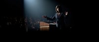 Professional businessman giving keynote speech at corporate conference with dramatic stage lighting and engaged audience in dark auditorium setting.