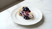 Creamy blueberry cheesecake slice topped with fresh berries on white plate, homemade dessert ready for cafe menu or bakery display.