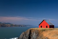 Red wooden barn perched dramatically on coastal cliff edge overlooking vast ocean bay and mountain ranges, golden grass swaying in foreground against deep blue sky.