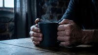 Senior man hands holding steaming hot coffee mug in cozy indoor setting with natural window light creating warm atmospheric mood for relaxation concepts.