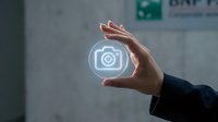 Hand holding glowing camera icon in modern office environment representing digital photography services and visual content creation for business.