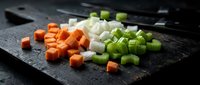 Fresh diced vegetables on dark cutting board with knife for cooking preparation. Colorful carrots, onions and celery ready for culinary use in kitchen.