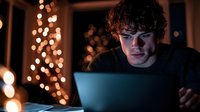 Young man with curly dark hair and fair complexion working on laptop at night, surrounded by warm golden bokeh lights, focused expression, indoor setting for tech, remote work, or online education concepts.