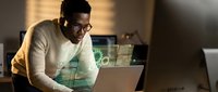 Young African American businessman working late at office desk with laptop computer in warm evening light wearing glasses and casual sweater.