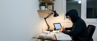 Young Middle Eastern man in black hoodie working late at night on laptop computer at modern home office desk with task lamp and books creating focused workspace atmosphere.