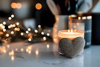 Glowing candle in glass holder decorated with silver glitter heart against blurred bokeh lights creating romantic atmosphere on marble surface.