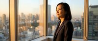 Asian woman in dark business blazer standing by floor-to-ceiling windows overlooking urban cityscape at golden hour, contemplative professional in modern office setting.
