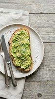 Avocado toast on sourdough bread with seeds and herbs served on white plate with knife and fork on rustic wooden table background for healthy breakfast.