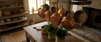 Rustic kitchen interior with copper pots hanging from rack above wooden table with fresh herbs and natural sunlight streaming through window.