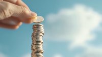 Stack of silver coins with hand placing another coin on top against blue sky with white clouds, representing financial growth, savings and investment concept.