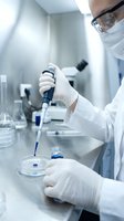 Male scientist in white lab coat and safety glasses using pipette in sterile laboratory conducting research experiment with petri dish and glassware.
