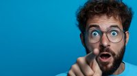 Young bearded man with glasses showing surprised expression and pointing finger at camera against blue background. Emotional portrait concept.