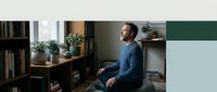 Middle-aged man with light skin tone and beard sitting on floor against wall in modern home office with bookshelves, plants, and natural light for remote work and lifestyle content.