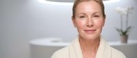 Middle-aged Caucasian woman in white bathrobe smiling in modern bathroom spa setting for wellness and self-care lifestyle concepts.