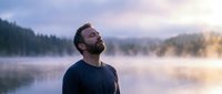 Peaceful bearded man looking up at sky during misty sunrise by mountain lake. Meditation, mindfulness and mental wellness concept for relaxation content.