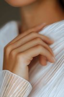 Delicate feminine hand with manicured nails gently touching white pleated fabric, creating soft and romantic mood for fashion and beauty content.