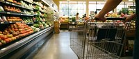 Shopper pushing cart through grocery store produce section with fresh fruits and vegetables displayed on shelves and refrigerated cases for retail shopping.
