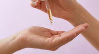 Woman applying golden serum oil from dropper bottle onto palm for skincare routine against soft pink background.