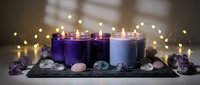 Purple and blue candles with crystals and gemstones on dark slate board surrounded by warm fairy lights creating peaceful meditation atmosphere.