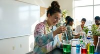 Young female scientist in tie-dye lab coat conducting chemistry experiment with colorful solutions in modern laboratory classroom setting.