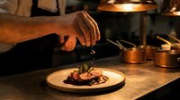 Professional chef garnishing gourmet meat dish with herbs in restaurant kitchen. Culinary artistry and fine dining preparation with warm ambient lighting.