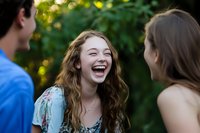 Young woman with wavy brown hair laughing joyfully while socializing with friends outdoors in natural setting with green bokeh background. Genuine happiness and connection.
