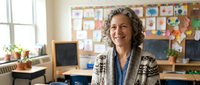 Smiling mature Caucasian female teacher with curly gray hair standing confidently in colorful elementary school classroom with student artwork displayed on walls.