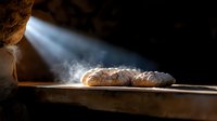 Fresh baked artisan bread loaf steaming on wooden surface with dramatic ray of light streaming through darkness, creating atmospheric food scene.