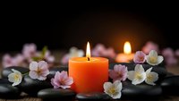 Orange candle burning with soft flame surrounded by delicate pink and white cherry blossoms on smooth black spa stones creating peaceful zen atmosphere.