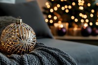 Glowing golden Christmas ornament with intricate snowflake pattern resting on cozy gray knit blanket against blurred festive lights and decorations background.