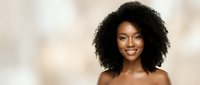 Young African American woman with natural curly hair smiling warmly at camera against soft beige background for beauty and skincare campaigns.