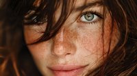 Young Caucasian woman with freckles and green eyes looking at camera with tousled brown hair covering part of face in natural portrait photography.