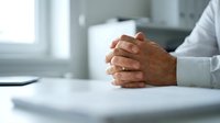 Closeup of mature male hands clasped together resting on a white desk, conveying confidence, patience, and professionalism in a business or medical setting.