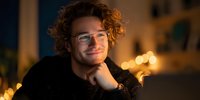 Young man with curly hair and glasses smiling at laptop in evening setting with warm bokeh lights in background.