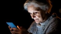 Senior Caucasian woman with gray hair using smartphone in dark room with warm lighting, elderly person browsing mobile device, technology and aging concept.