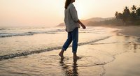 Woman walking barefoot on tropical beach at sunset with golden light reflecting on wet sand and gentle waves washing shore creating peaceful vacation moment.