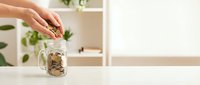 Woman hand putting coins into glass jar on white shelf for savings and financial planning goals.