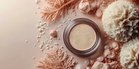 Luxury face powder in metallic container surrounded by delicate dried flowers and pearls on peachy pink background, flat lay cosmetic composition.