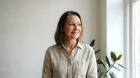Middle-aged woman with long dark hair and fair complexion, wearing a beige linen shirt, smiling while standing near a window with potted plant, natural daylight portrait for lifestyle and wellness content.