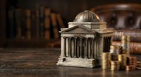 Miniature classical bank building model with columns and dome surrounded by golden coins on wooden surface with blurred books background for finance concepts.