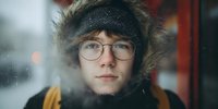 Young man in winter fur hood and glasses looking through foggy window on cold day, creating atmospheric portrait with condensation effect.