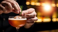 Professional bartender garnishing craft cocktail with fresh rosemary sprig in elegant coupe glass at upscale bar with warm ambient lighting.