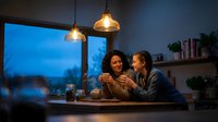 Cozy evening scene with young couple enjoying hot drinks together at home kitchen counter with warm pendant lighting and blue hour window view.