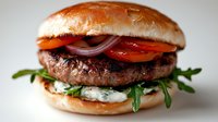 Juicy grilled beef burger with fresh vegetables, arugula and creamy sauce on brioche bun against light background, close up view of gourmet sandwich.