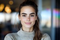 Young Caucasian woman with natural makeup and ponytail wearing beige sweater smiling at camera. Soft bokeh background creates professional portrait atmosphere.