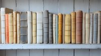 Row of vintage hardcover books with worn fabric spines in neutral beige, cream, and coral tones arranged on white wooden shelf, creating nostalgic library atmosphere.