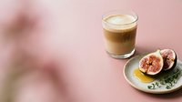 Layered coffee drink with creamy foam served alongside fresh figs and honey on ceramic plate against soft pink background for café and food styling.