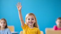 Cheerful elementary school student raising hand to answer question in classroom. Blonde girl in yellow t-shirt showing enthusiasm during lesson.