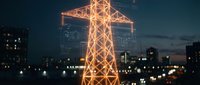 Illuminated electrical transmission tower glowing against urban skyline at dusk with city lights and modern buildings creating dramatic infrastructure scene.